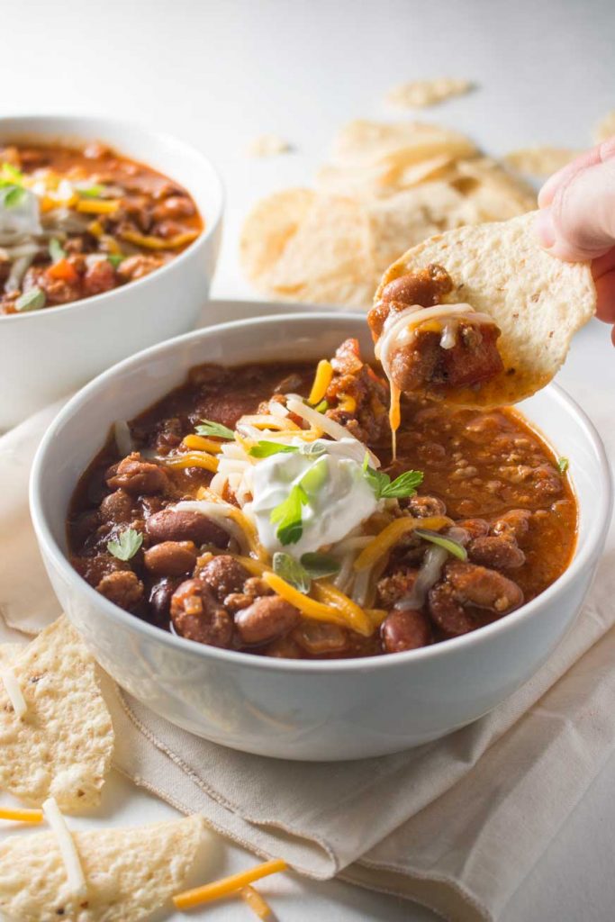 Hand holding tortilla chip scooping stovetop turkey chili from white bowl with sour cream