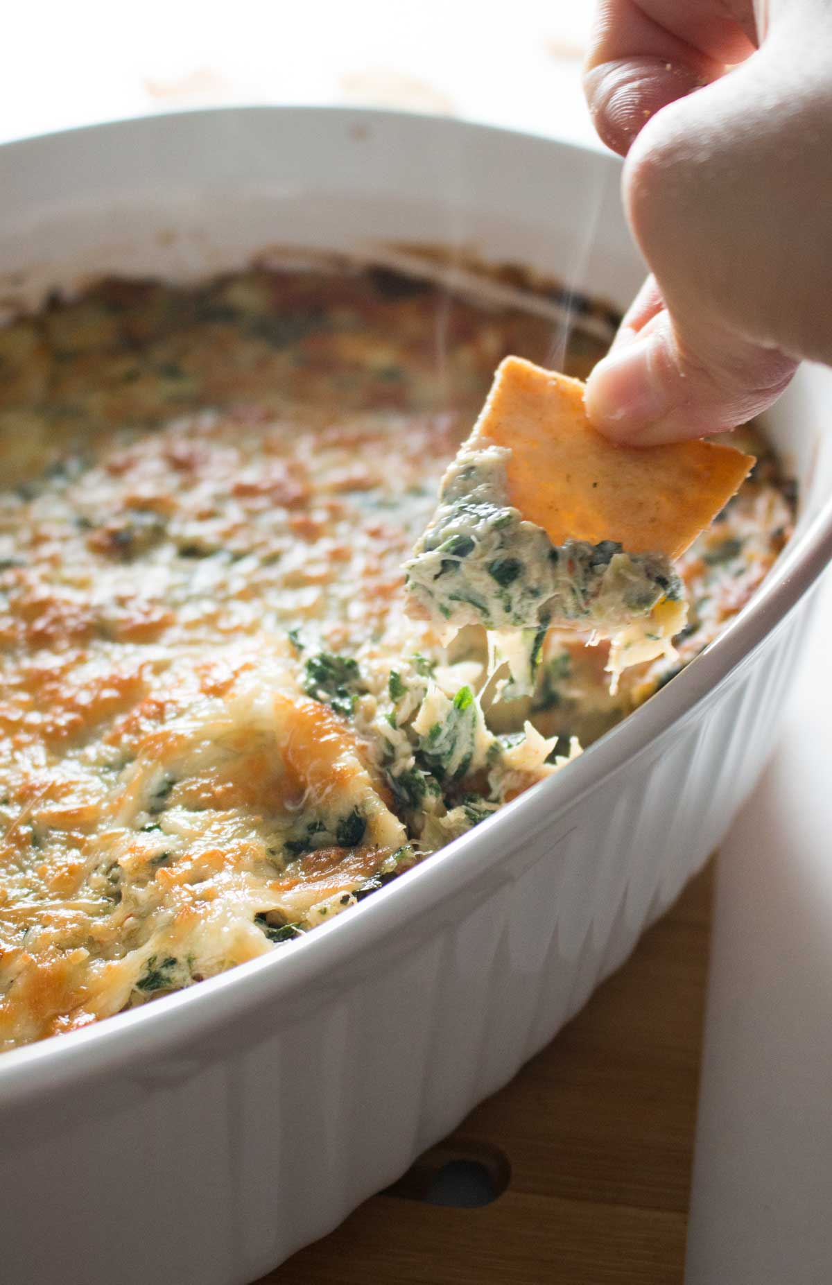 Closeup of pita chip scooping steaming baked spinach, artichoke, and crab dip in white casserole dish