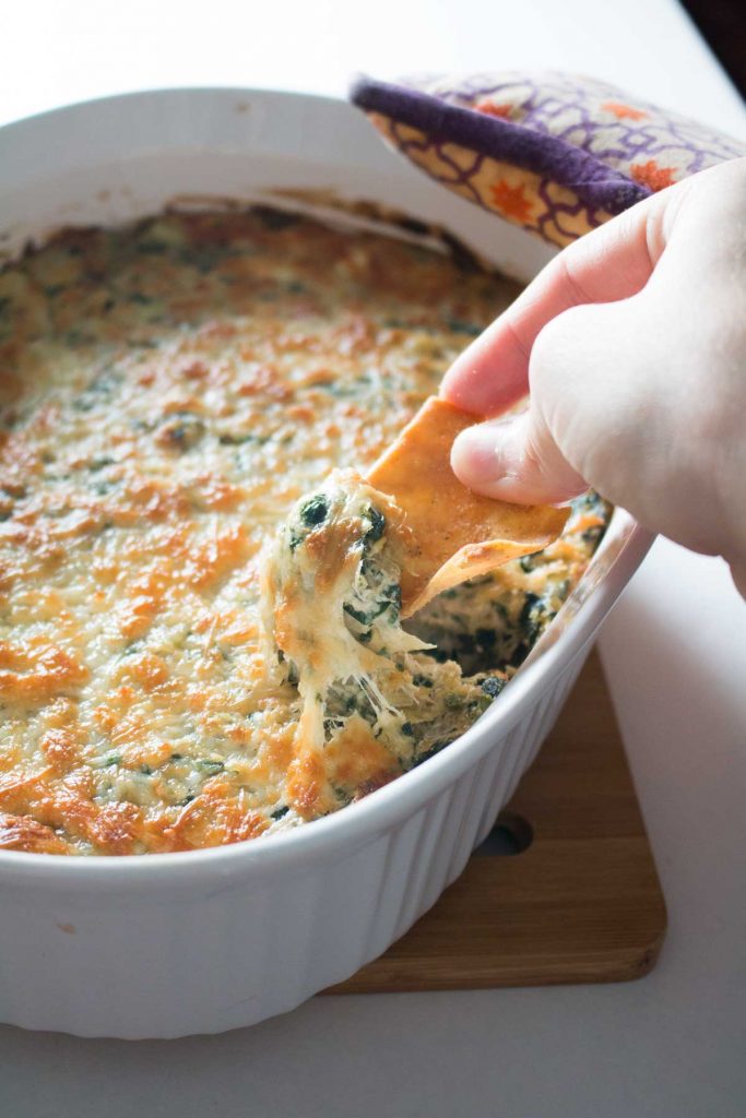 Hand holding pita chip scooping cheesy baked spinach, artichoke, and crab dip in white casserole dish