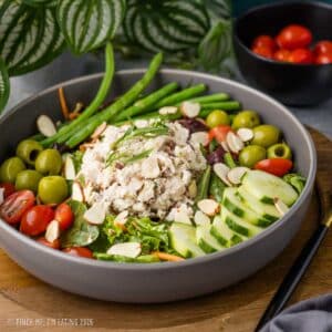 Eye-level view of a French bistro salad in a gray ceramic bowl topped with a scoop of tarragon chicken salad, blanched green beans, sliced cucumbers, grape tomatoes, green olives, and sliced almonds.