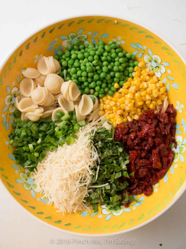 Ingredients for sun-dried tomato pasta salad with basil, corn, peas, and parmesan in a yellow flowered bowl.