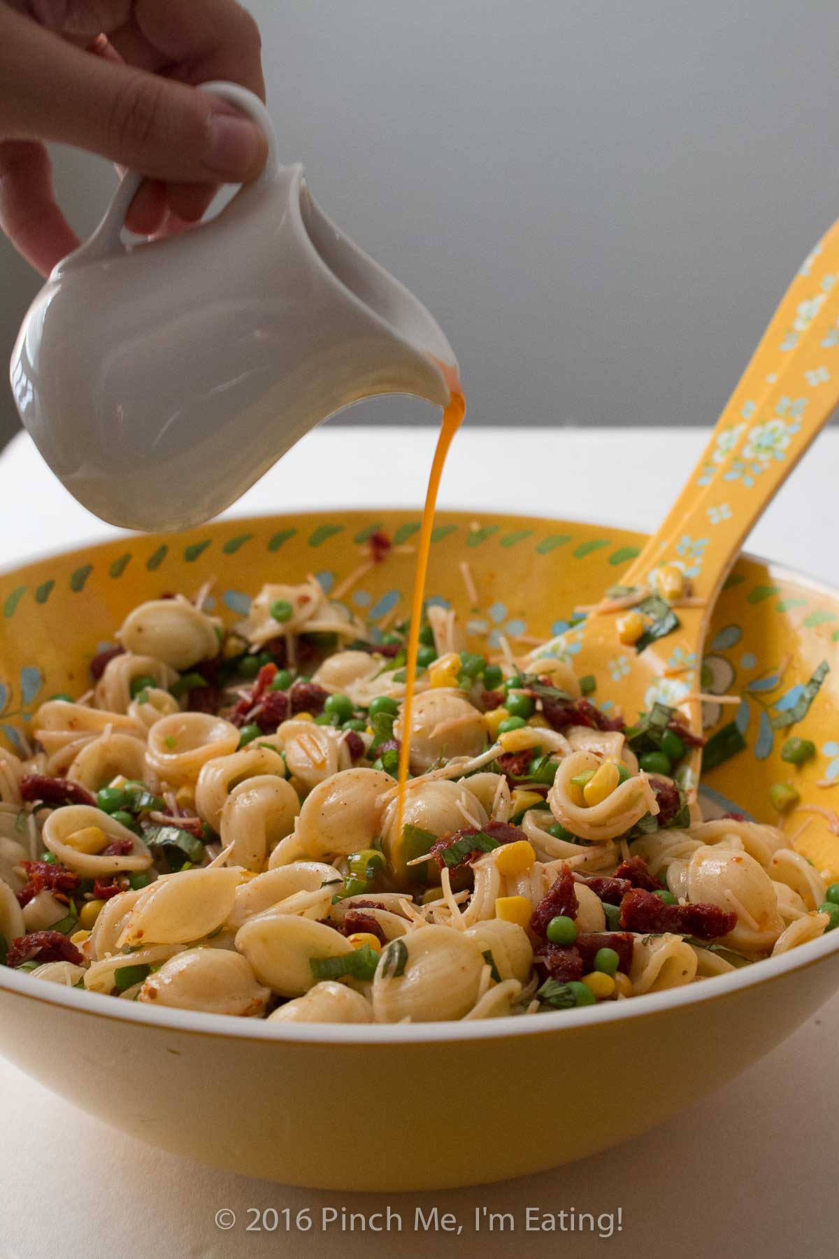 Drizzling sun-dried tomato vinaigrette out of a white pitcher onto orecchiette pasta salad.