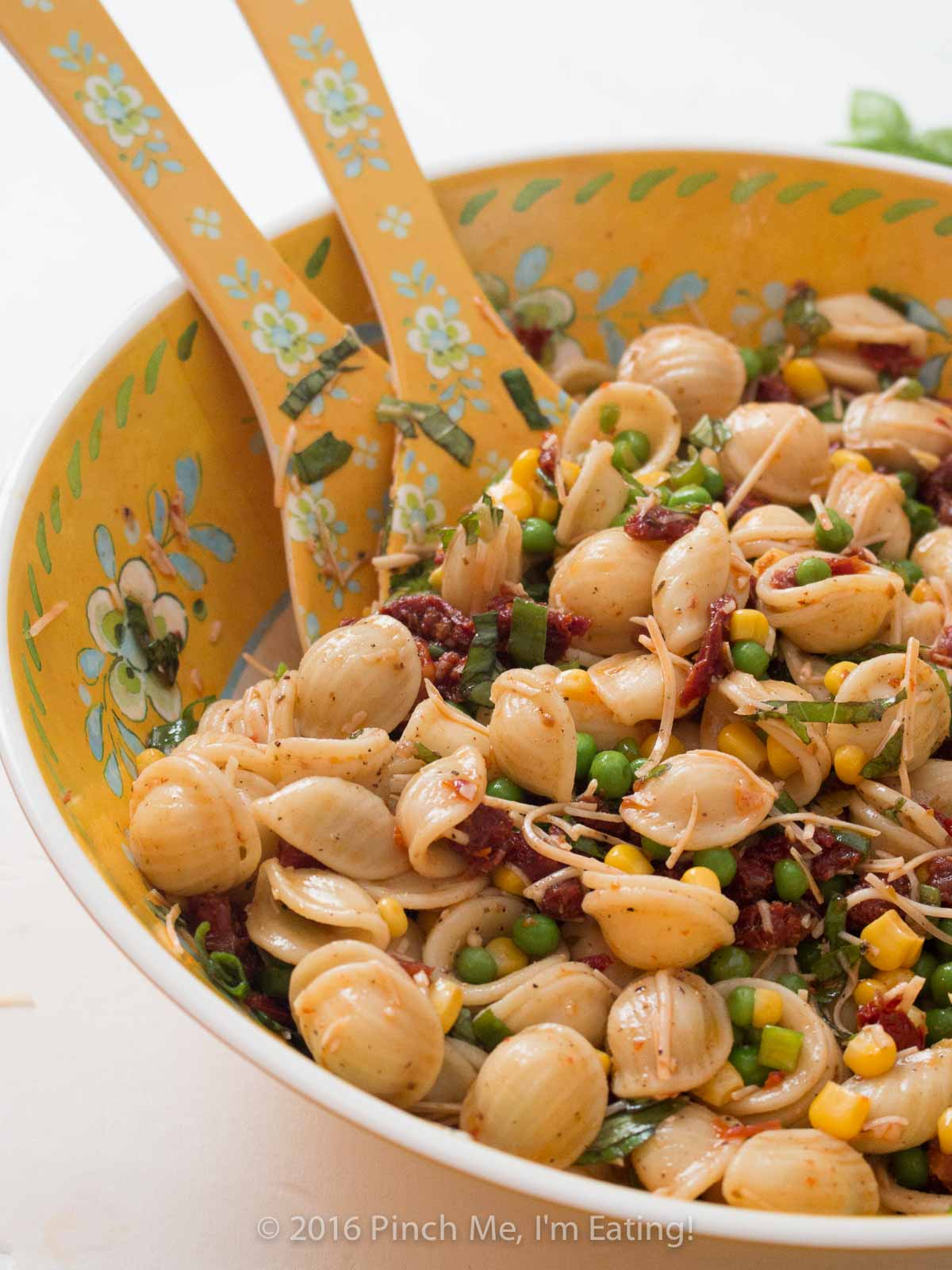 Sun-dried tomato orecchiette pasta salad with basil, corn, peas, and parmesan in a yellow flowered bowl.