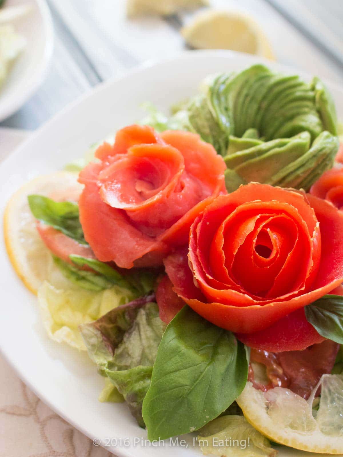 Smoked salmon, tomato, and avocado roses with salad on a white plate.