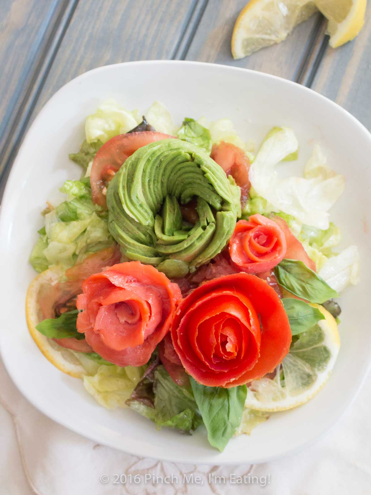 Smoked salmon, tomato, and avocado roses with salad on a white plate.