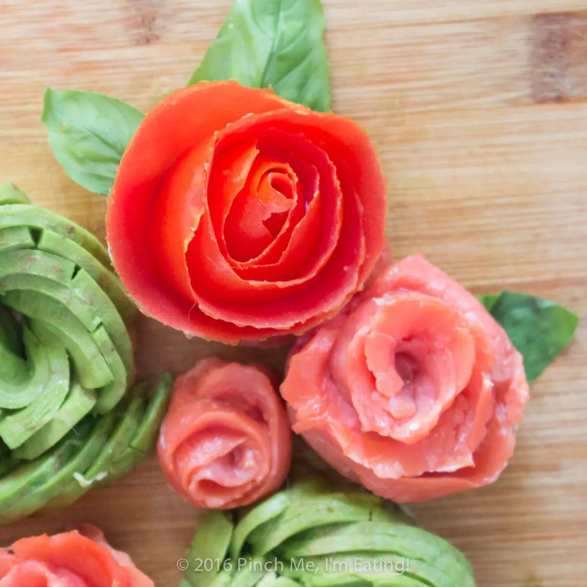 Decorative roses made from tomato, avocado, and smoked salmon with basil leaves on a wooden cutting board. 