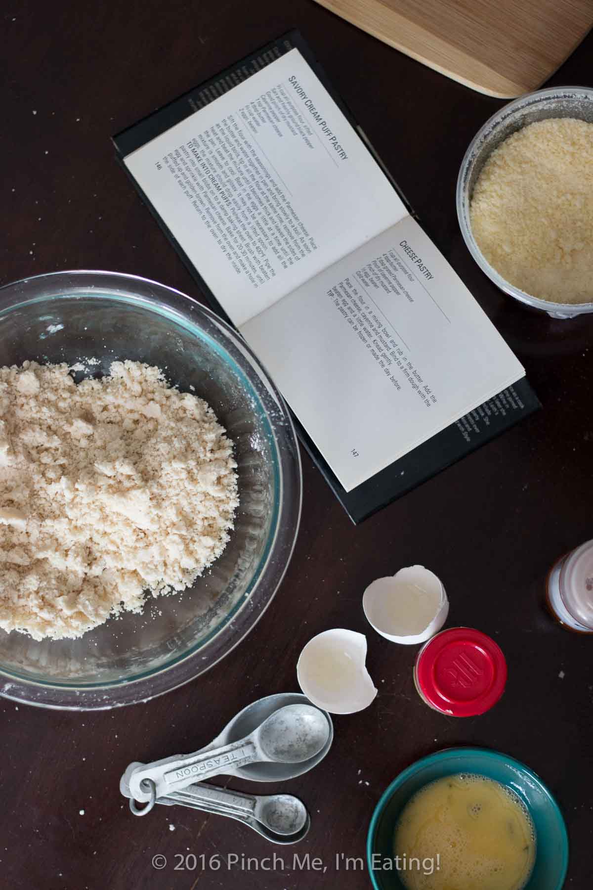 Overhead shot of pastry dough in a glass bowl, grated cheese, egg and eggshell, and a small open cookbook on a wooden table.