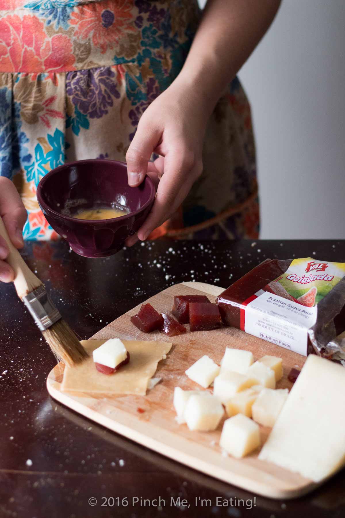 Woman in floral apron brushing egg onto guava and manchego cheese pastry with a pastry brush.