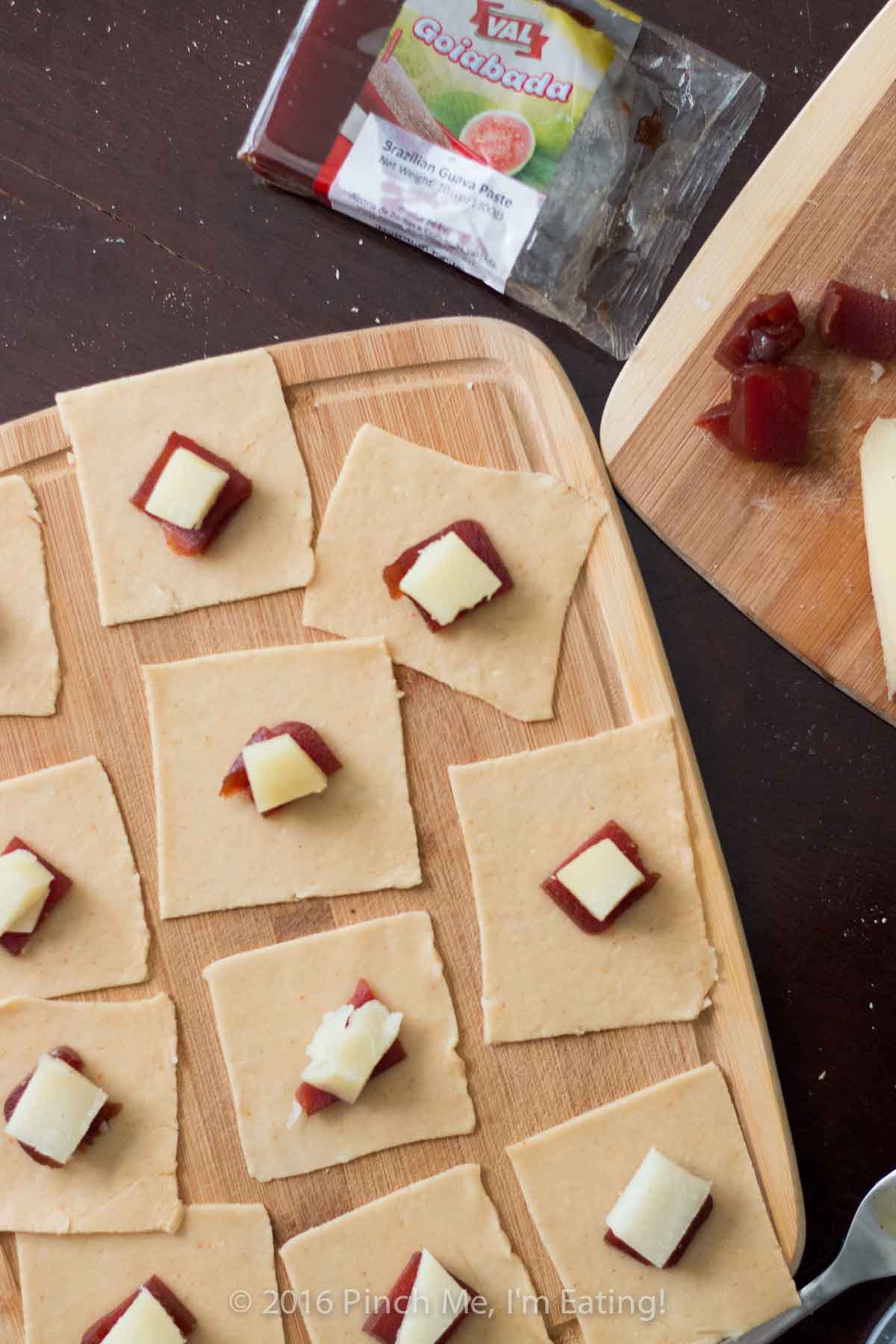 Squares of pastry dough topped with pieces of guava paste and manchego cheese on a wooden cutting board.