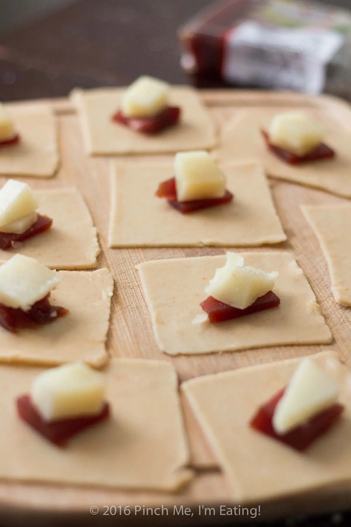 Squares of pastry dough topped with pieces of guava paste and manchego cheese on a wooden cutting board.