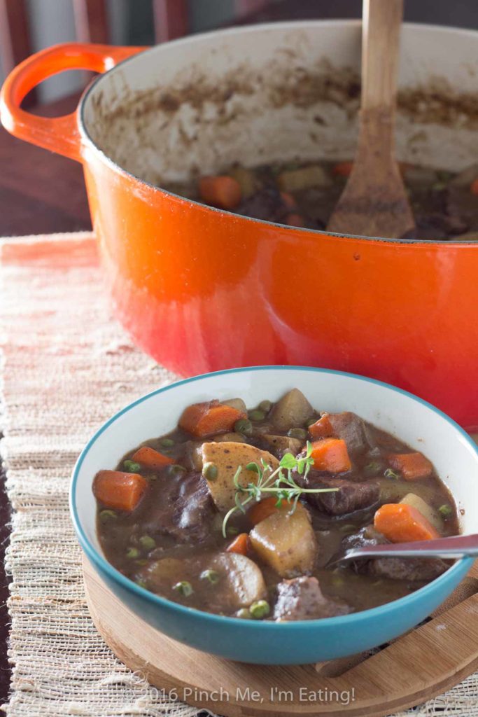 Stove top beef stew with red wine with sprig of thyme in blue bowl next to Dutch oven