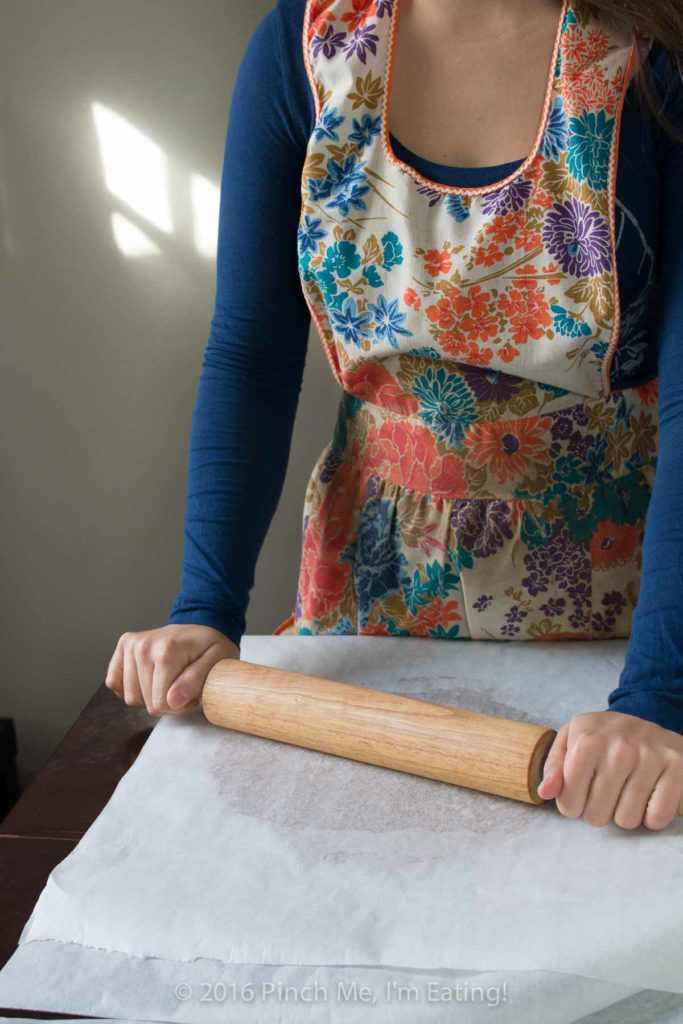 Woman with rolling pin in vintage flowered apron, rolling cookie dough between sheets of parchment paper