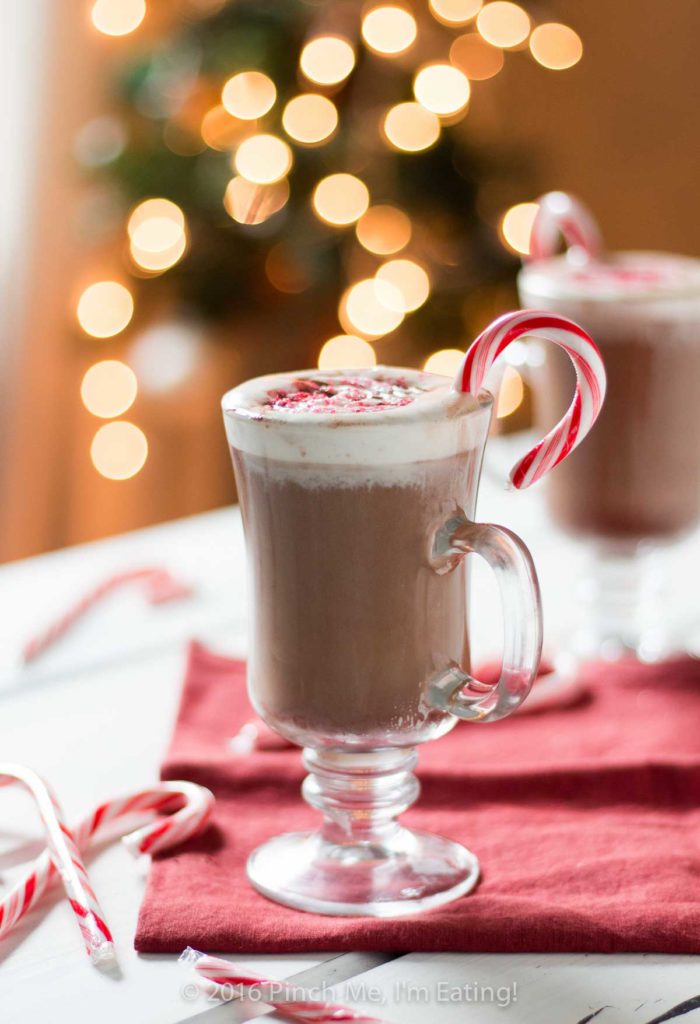 Peppermint mocha in clear glass mug with whipped cream and candy canes on red napkin. Blurred Christmas lights are in the background.