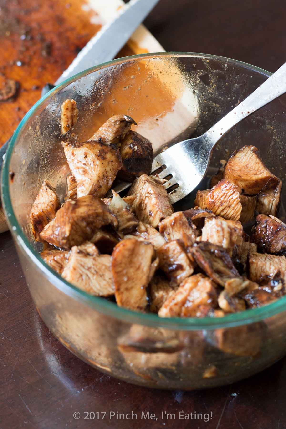 Cooked chicken chunks marinating in balsamic vinegar in a clear bowl with a fork.
