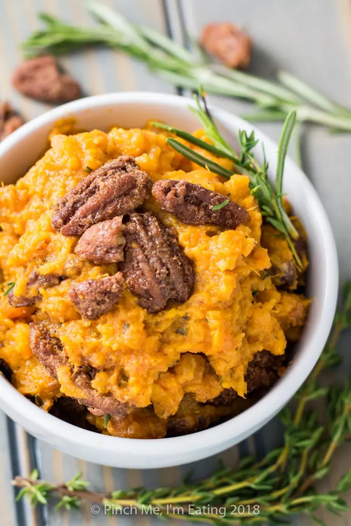 Closeup of rosemary mashed sweet potatoes with pecans candied in cinnamon sugar in white bowl, with sprig of fresh rosemary