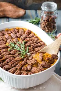 Spoon serving sweet potato casserole from white serving dish, topped with candied pecans and rosemary