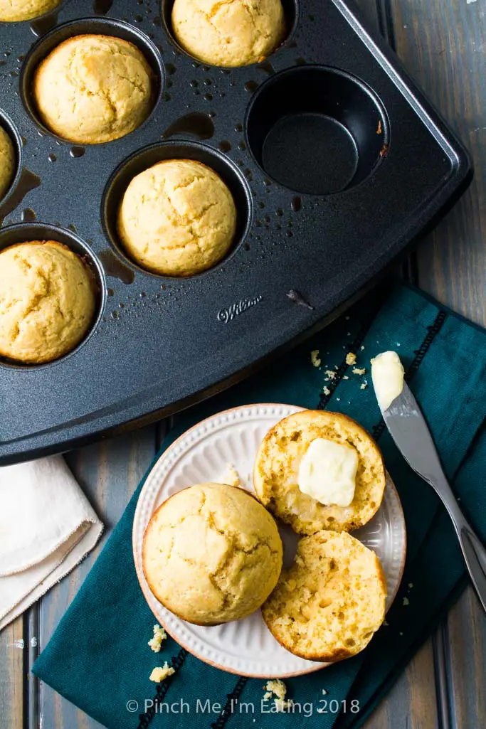 A cornbread muffin split open and buttered on a plate next to a muffin tin full of cornbread muffins