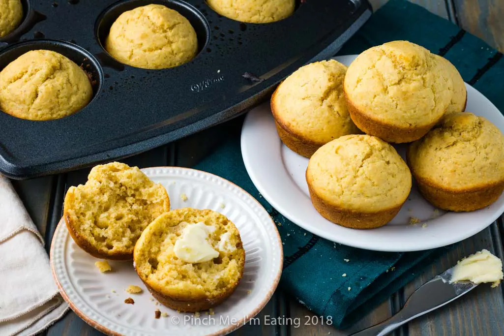 A cornbread muffin split open with butter, next to a white plate piled with cornbread muffins
