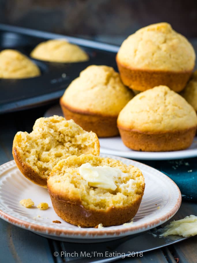 A cornbread muffin split open with butter in front of a white plate of cornbread muffins