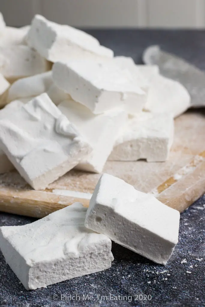 A pile of homemade marshmallows on a cutting board with two marshmallows on a surface in front