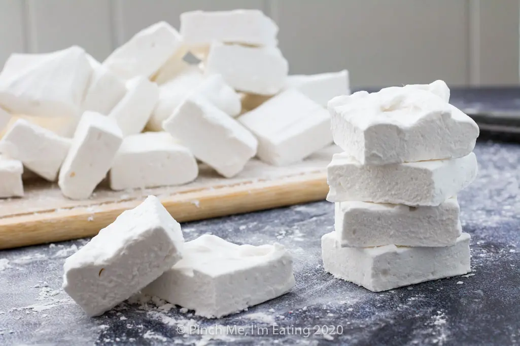 Several homemade marshmallows are stacked on a surface in front of a pile of marshmallows on a cutting board.