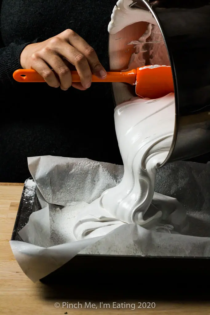 A spatula scrapes homemade marshmallow mixture into a baking pan lined with parchment paper