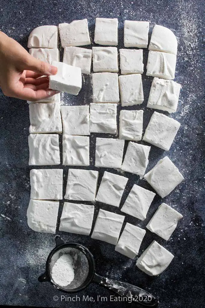 A hand picks up a homemade marshmallow. More marshmallow squares are arranged on a surface.