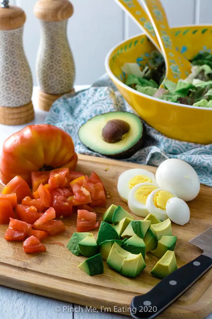 Chopped tomato and avocado and sliced hard boiled egg on cutting board in front of large salad bowl