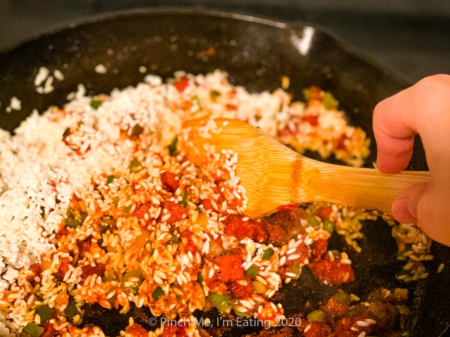 A wooden spatula stirring uncooked white rice in a cast iron skillet with veggies, sausage, and tomato paste
