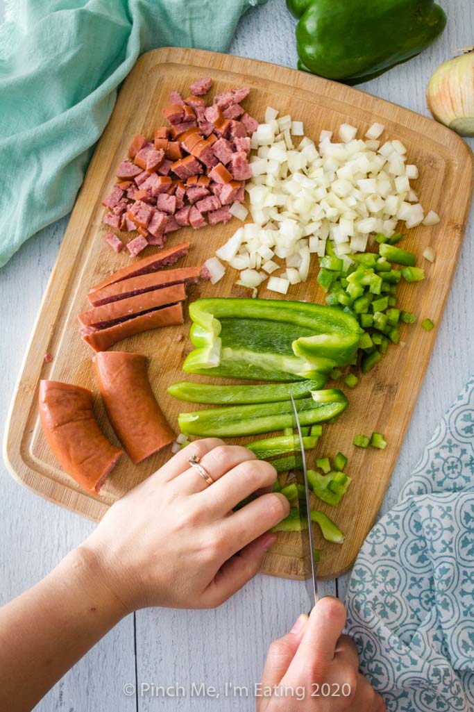 A hand with knife cuts green bell peppers on a wooden cutting board with smoked sausage and diced onions