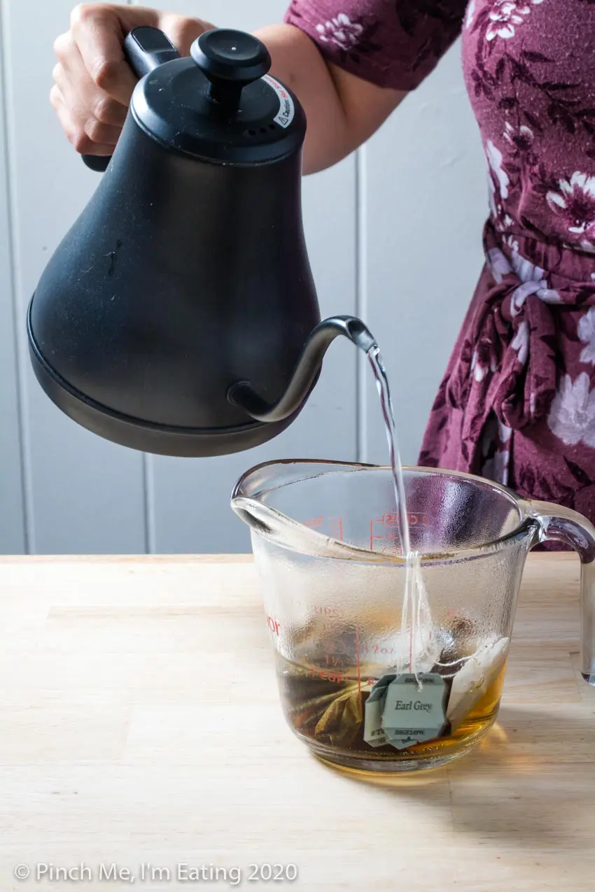 A woman pouring water from an electric kettle into a measuring cup with four Earl Grey tea bags