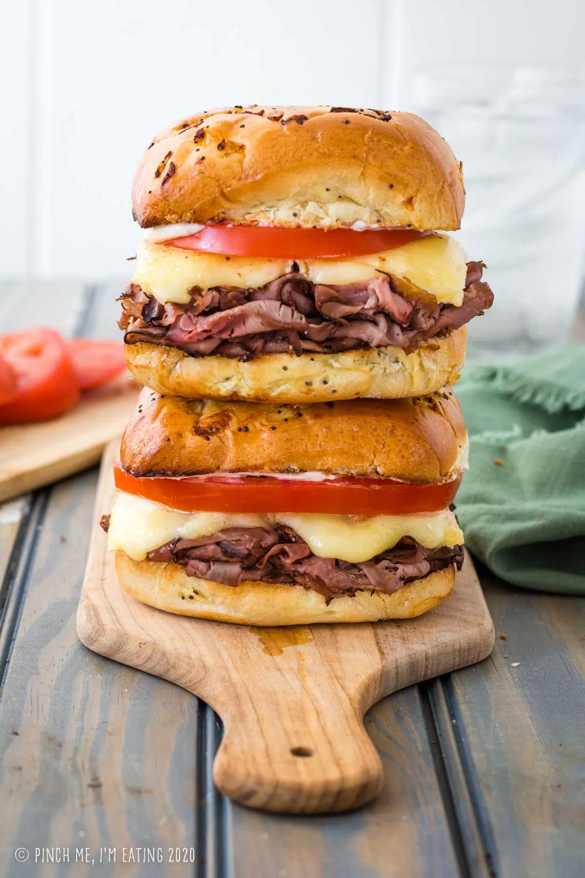 Two hot roast beef and brie sandwiches with tomato stacked on a wooden cutting board with tomato and napkin in the background.