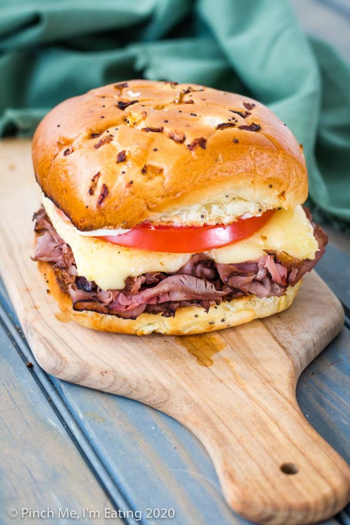 Roast beef sandwich with melted brie and tomato on an onion roll on a wooden cutting board with green napkin in the background