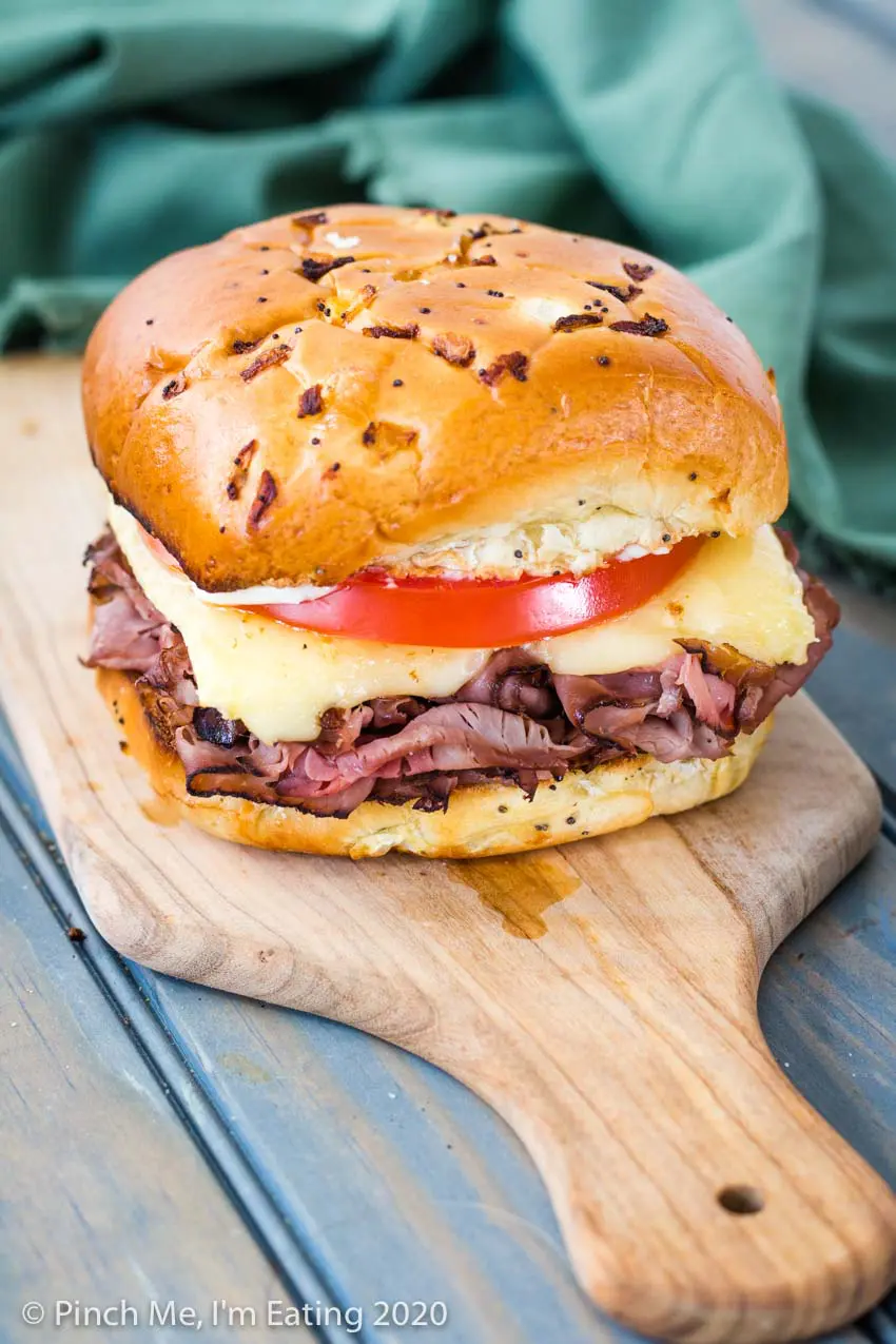 Roast beef sandwich with melted brie and tomato on an onion roll on a wooden cutting board with green napkin in the background