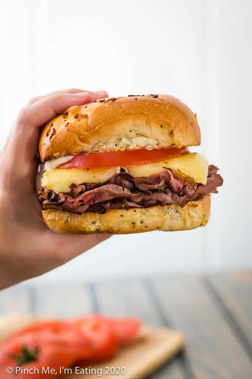 Hand holding roast beef and brie sandwich with tomato on an onion roll in front of a white background