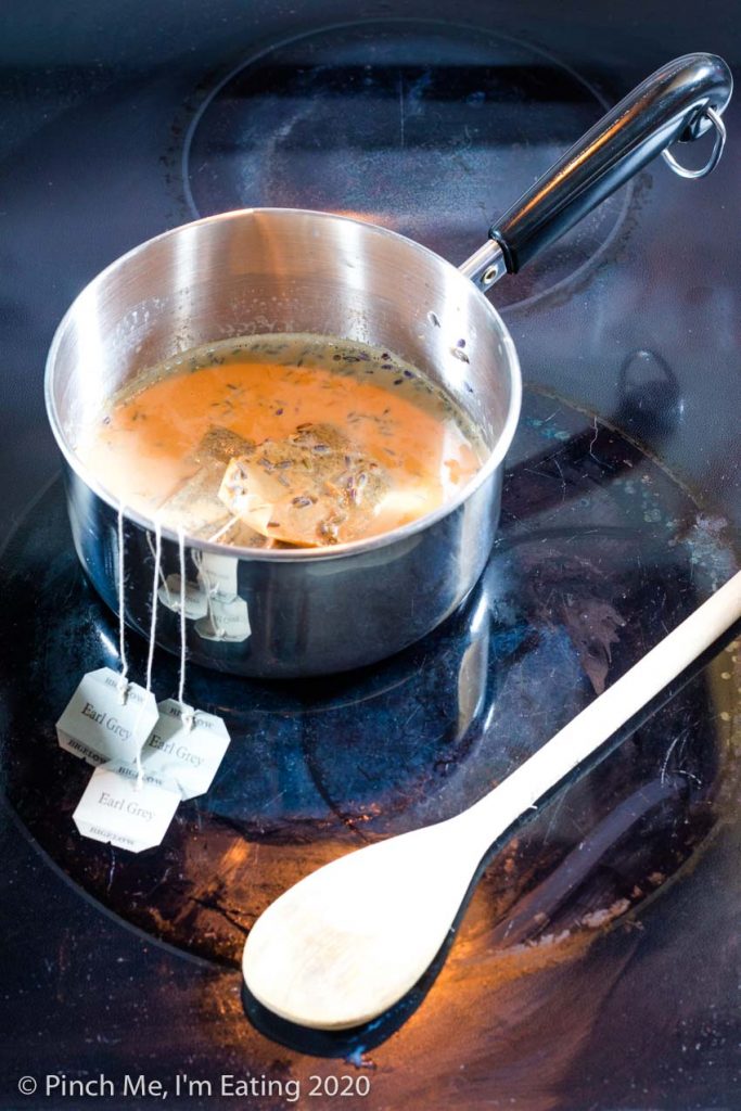 A small saucepan of simmering milk is steeping with three Earl Grey teabags and dried lavender flowers.