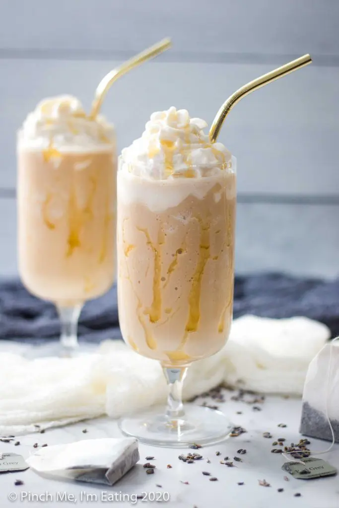 Two glasses drizzled with honey are filled with lavender Earl Grey Frappuccinos and whipped cream, with gold straws. Tea bags and dried lavender are in the foreground.