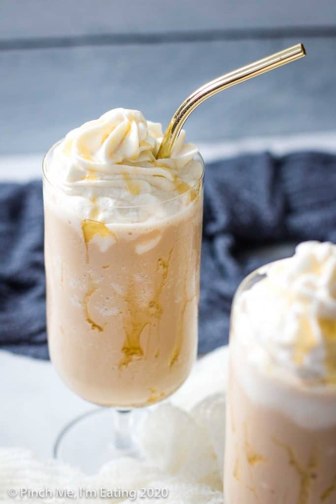 A glass drizzled with honey is filled with a lavender Earl Grey Frappuccino and whipped cream, with a gold straw. A second glass is blurred in the foreground.
