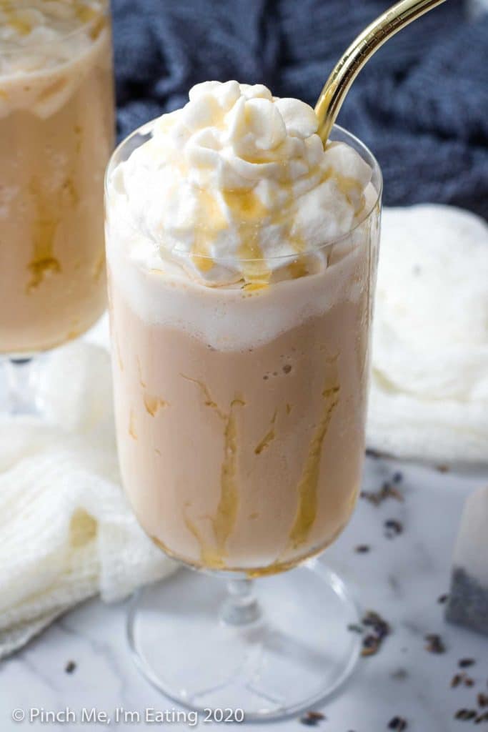 A glass drizzled with honey is filled with a lavender Earl Grey Frappuccino and whipped cream, with a gold straw. A second glass is blurred in the background.