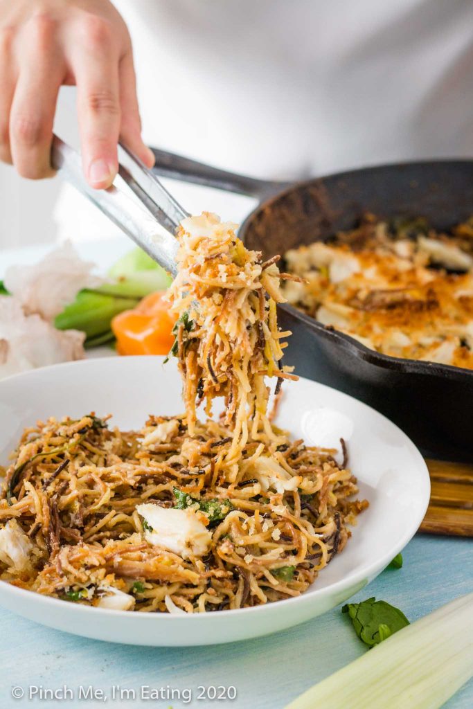 Tongs serving toasted angel hair pasta with crab into a white bowl, with a cast iron skillet in the background