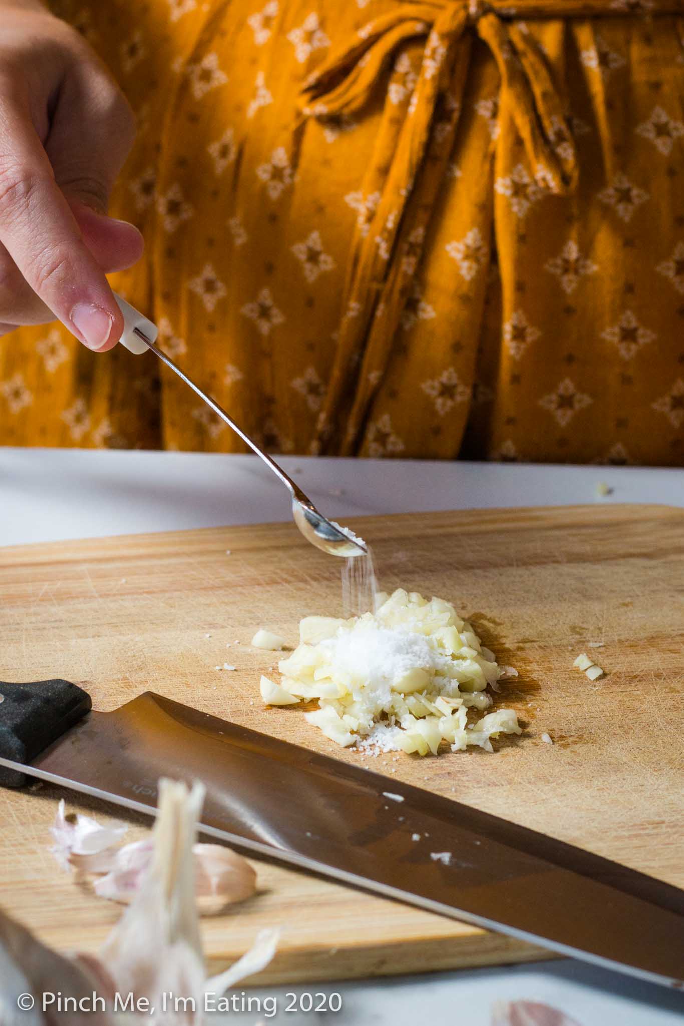Hand pouring kosher salt onto pile of minced garlic on wooden cutting board