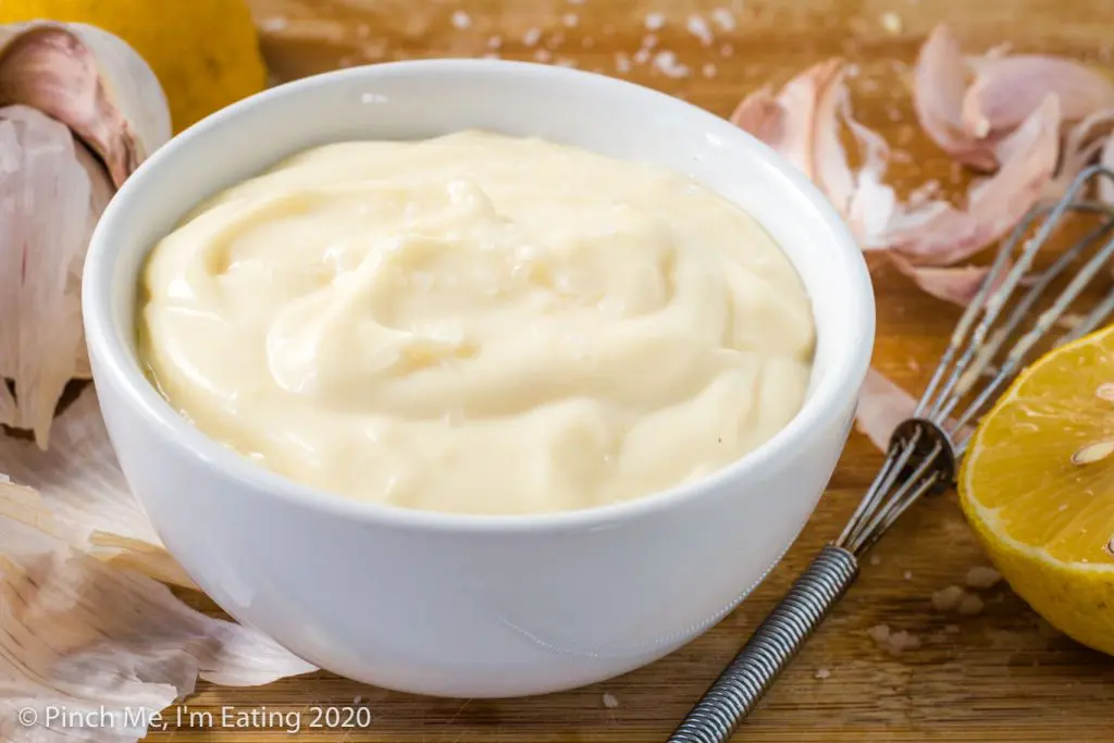 Easy garlic aioli in a small white bowl with a head of garlic, lemon, and whisk to the side