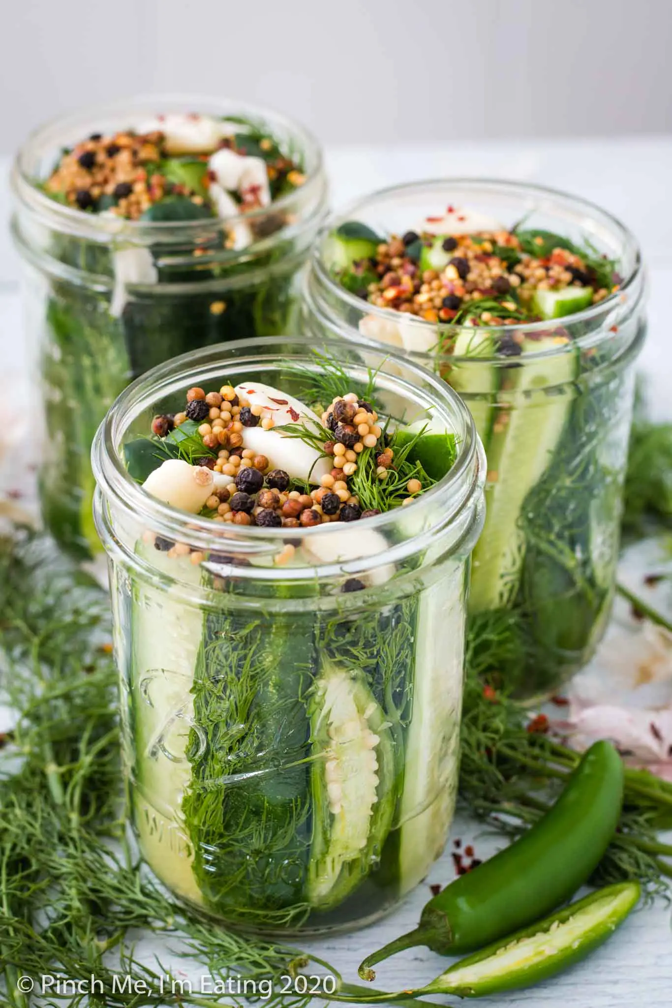 Three jars of spicy refrigerator dill pickles with fresh dill and serrano peppers in foreground. Jars are topped with garlic and pickling spices.