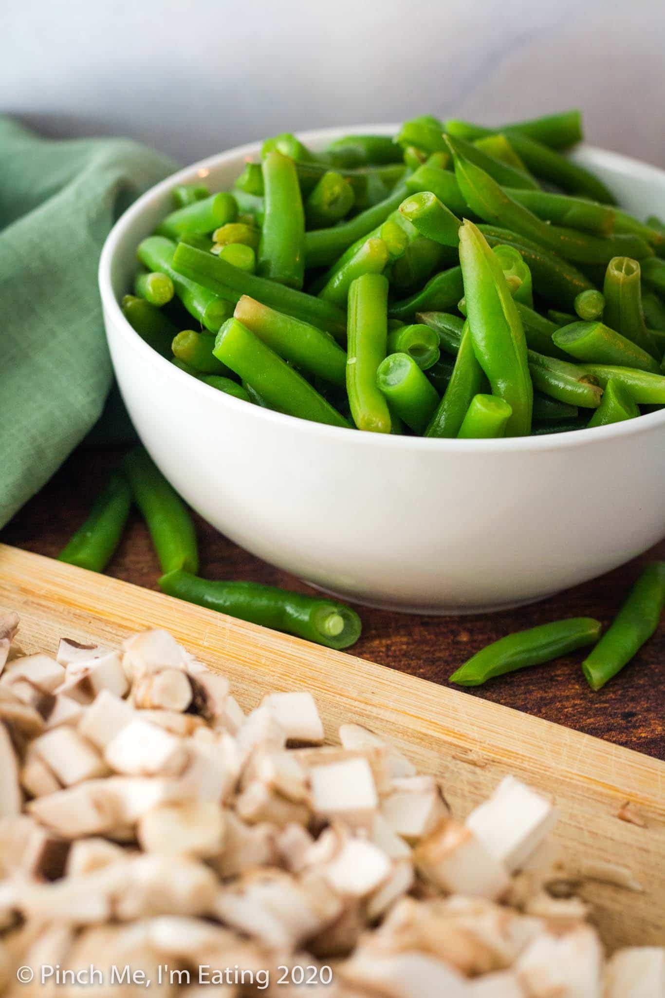 A white bowl of fresh blanched green beans with blurred mushrooms in the foreground