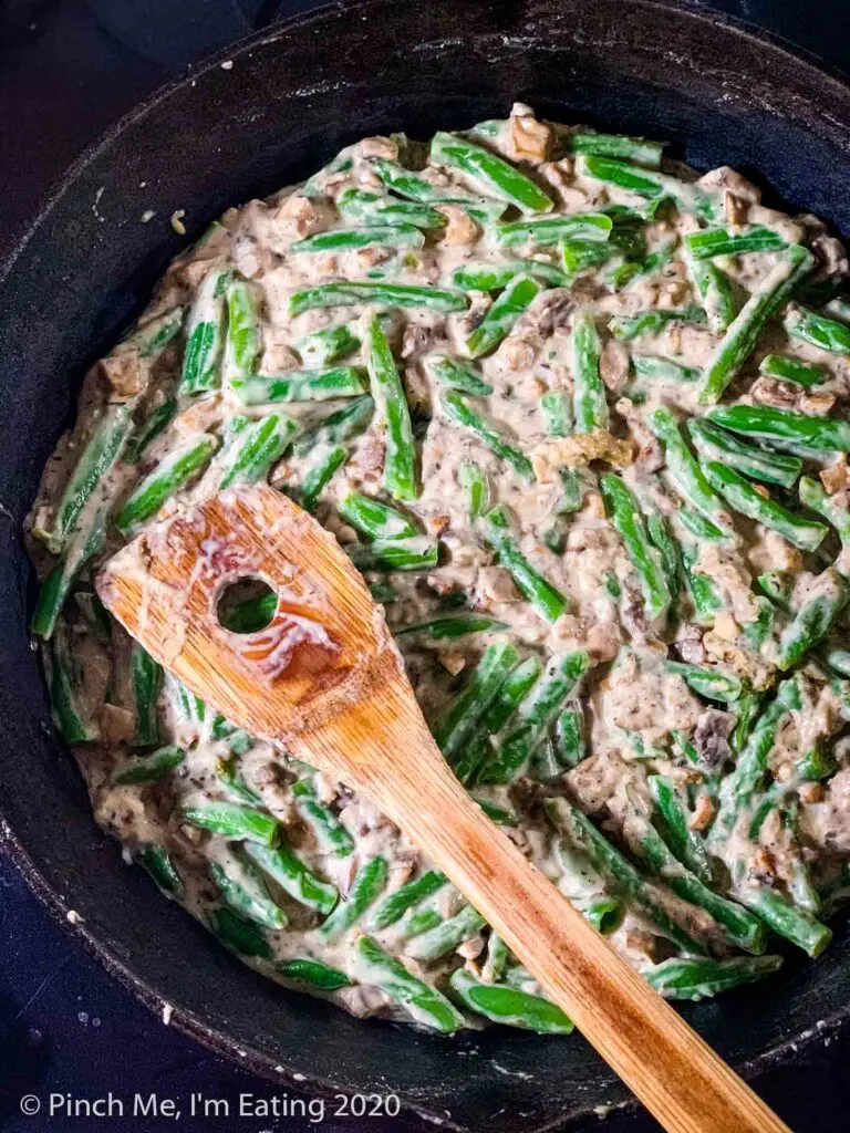 Fresh green beans mixed into homemade cream of mushroom soup in a cast iron skillet with a wooden spoon