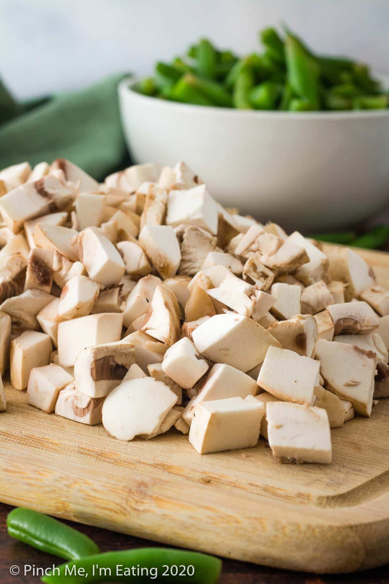 Bamboo cutting board with a pile of diced mushrooms on it, with a white bowl of fresh green beans blurred in the background