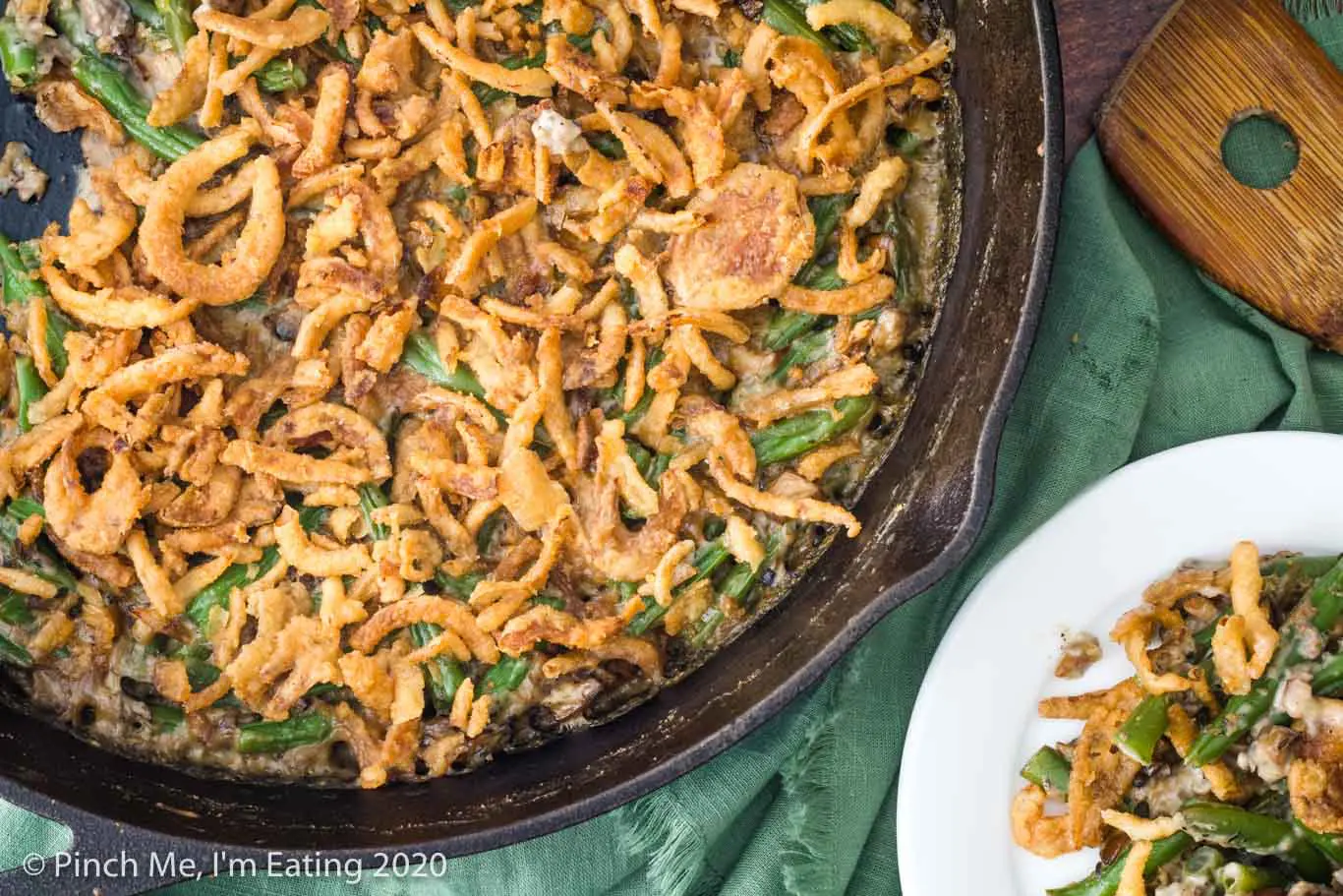 Overhead view of fresh green bean casserole in cast iron skillet with a single serving on a white plate and wooden spoon