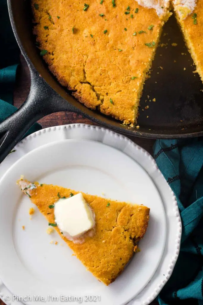 A wedge of cornbread with butter on top on a white plate next to a cast iron skillet of Southern buttermilk cornbread