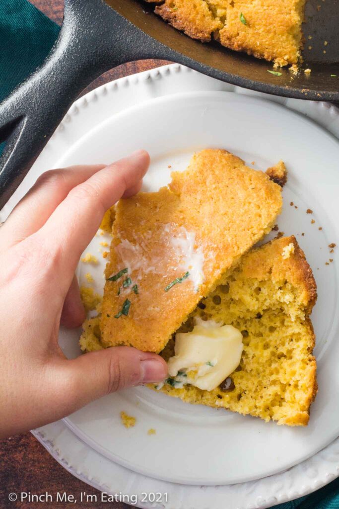 A hand picking up a wedge of Southern buttermilk cornbread split open on a white plate with butter on top