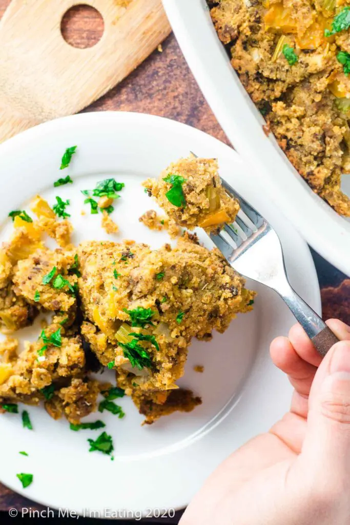 Overhead view of a hand holding a bite of Southern cornbread dressing on a fork above a serving on a small white plate